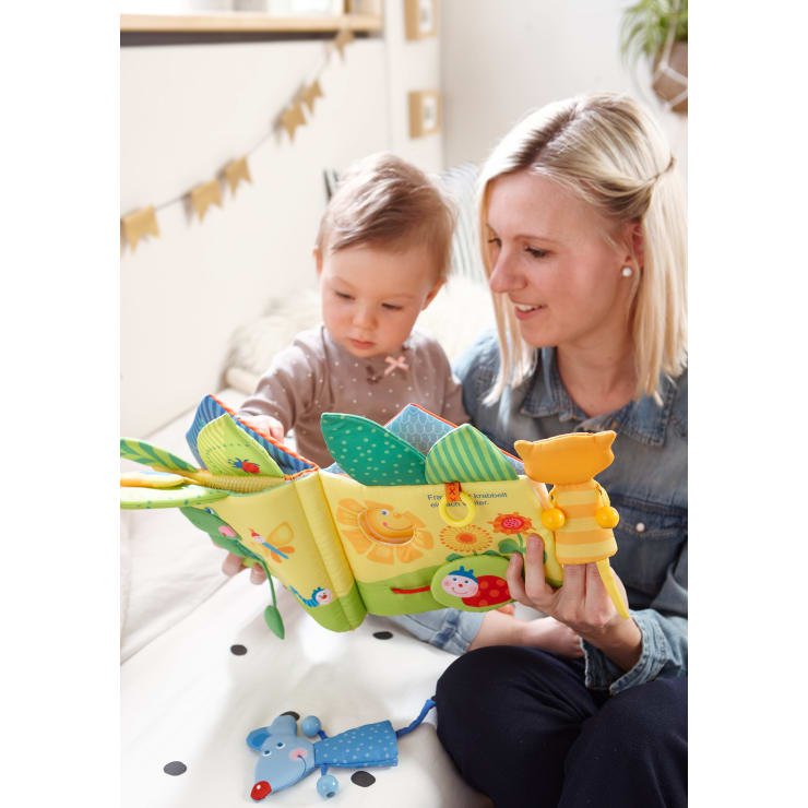 Eine Frau und ein kleines Kind sitzen eng beieinander auf einem Bett und betrachten ein buntes, weiches Stoffbuch mit Tierillustrationen und beigefügtem Spielzeug. Das Zimmer ist sanft beleuchtet und mit goldenen Wimpeln an der Wand geschmückt.