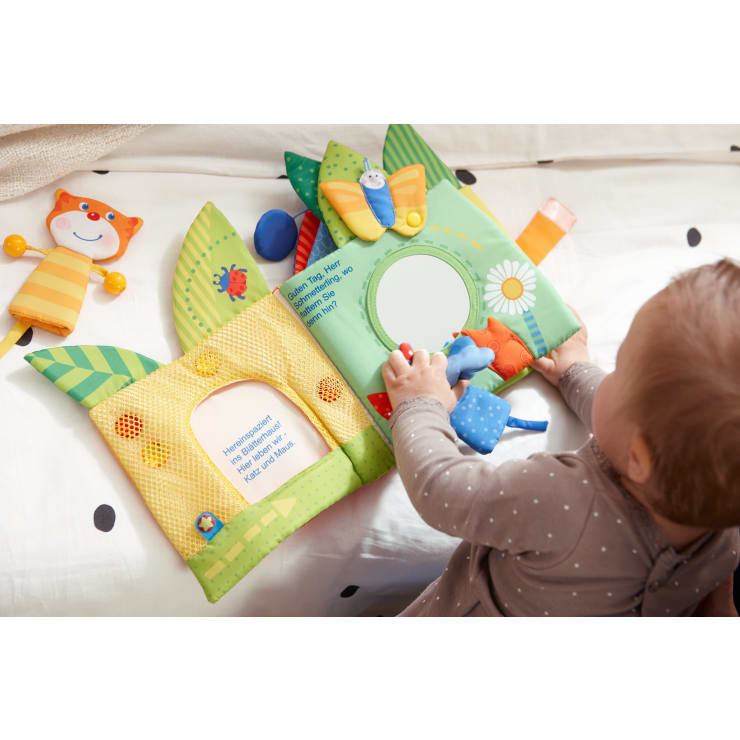 Ein Baby im grauen Polka-Dot-Outfit spielt mit einem farbenfrohen Buch aus weichem Stoff, das interaktive Elemente wie einen Spiegel, einen Schmetterling und kleine Stofftiere auf hellem Hintergrund enthält.