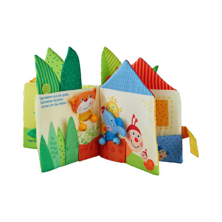 Ein farbenfrohes Stoff-Babybuch mit weichen, wattierten Seiten, die fröhliche Cartoon-Tiere, eine Sonne und deutschen Text zeigen, mit strukturierten grünen und blauen Akzenten entlang der Ränder.