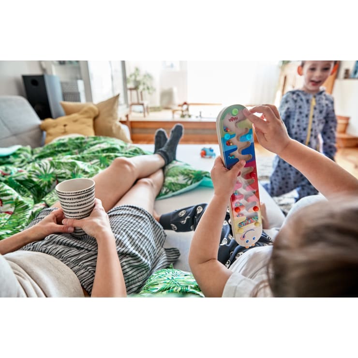 Ein Erwachsener sitzt auf dem Sofa und hält eine gestreifte Tasse in der Hand, während ein Kind in der Nähe mit einem tragbaren Marmor-Labyrinth-Spiel spielt; ein weiteres Kind steht im Hintergrund, alles in einem hell erleuchteten Wohnzimmer.