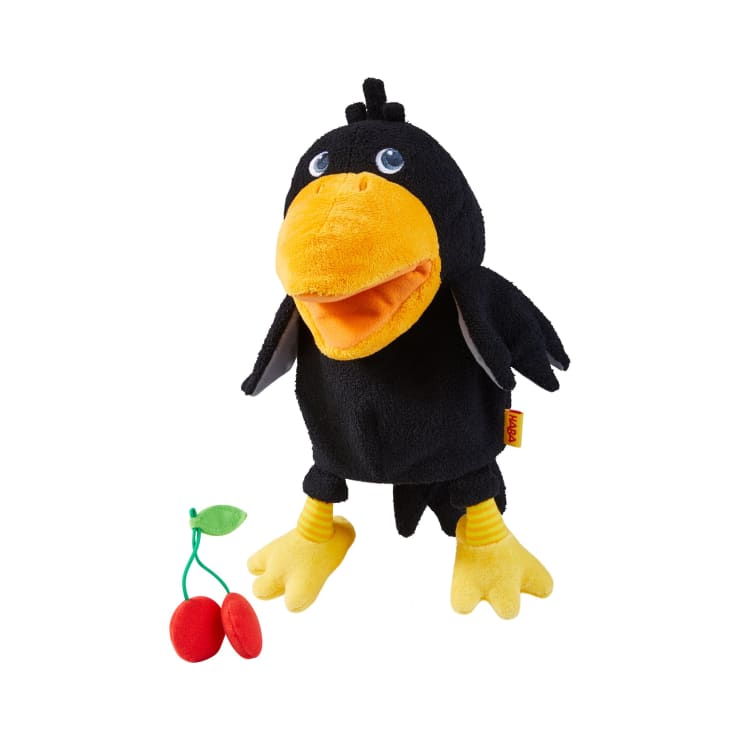 Eine schwarze Plüschkrähe mit gelben Füßen und Schnabel steht aufrecht neben einer kleinen Stoffkirsche mit zwei roten Kirschen und einem grünen Blatt, alles auf weißem Hintergrund.