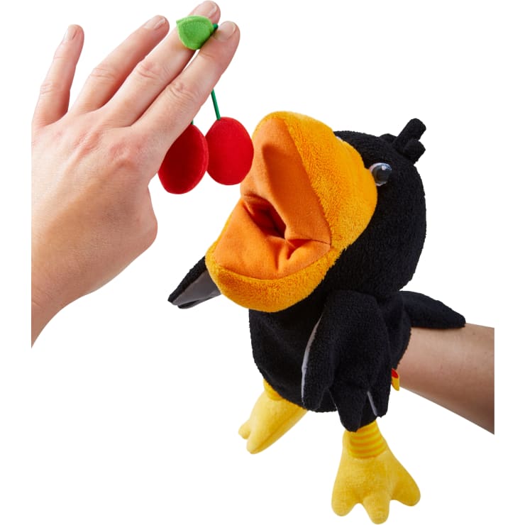 Eine Hand hält zwei rote Pom-Pom-Kirschen mit einem grünen Filzblatt vor einer schwarz-gelben Vogelpuppe mit offenem Maul, die von einer anderen Hand gesteuert wird, alles vor einem weißen Hintergrund.