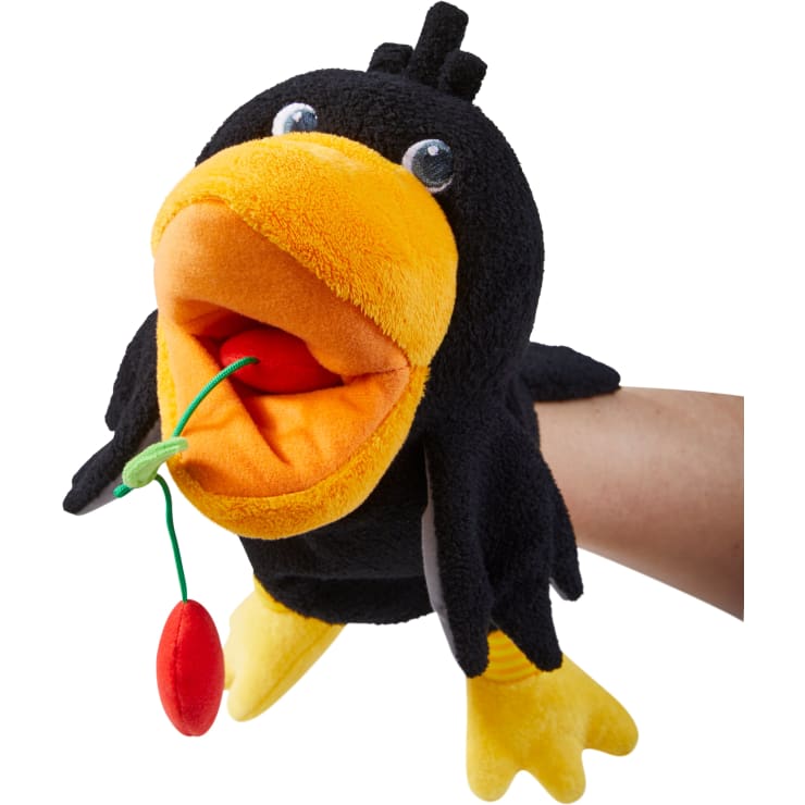 Eine schwarz-gelbe Vogelhandpuppe mit weit geöffnetem Schnabel hält ein rotes und grünes Stoffobjekt in ihrem Mund. In der Puppe, die vor einem weißen Hintergrund fotografiert wurde, befindet sich ein Personenarm.