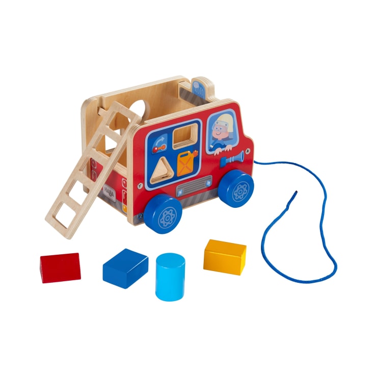 Ein hölzernes Spielzeug-Feuerwehrauto mit einer Leiter und einer Zugschnur hat an den Seiten Löcher zum Sortieren der Formen. Vier bunte Blöcke - ein rotes Quadrat, ein blaues Rechteck, ein blauer Zylinder und ein gelbes Rechteck - sind davor platziert.