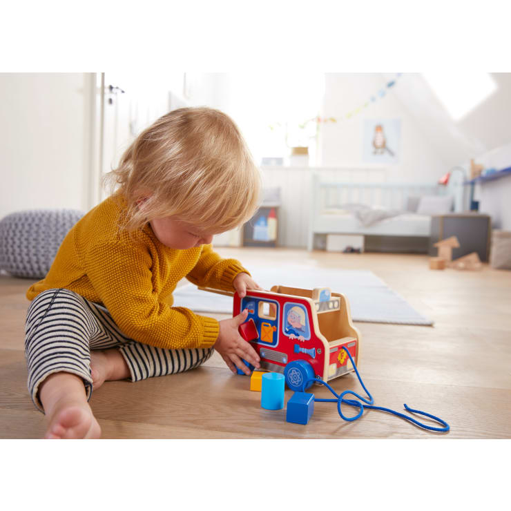 Ein kleines Kind in einem gelben Pullover sitzt auf einem Holzboden und spielt mit einem bunten Holzspielzeug, zu dem auch Formblöcke gehören, in einem hellen Raum mit hellem Dekor und einigen Spielzeugen im Hintergrund.
