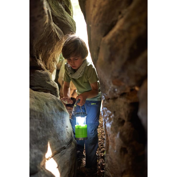 Ein Kind in Outdoor-Kleidung geht mit einer grünen Laterne in der Hand durch eine enge Felsspalte. Das Sonnenlicht dringt durch die Felsen und erhellt den Weg vor ihm.