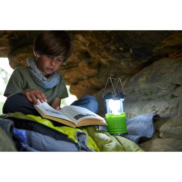 Ein kleiner Junge sitzt in einer Höhle oder einem Felsunterstand und liest ein Buch mit einer grünen Campinglaterne neben sich. Er ist umgeben von Schlafsäcken und Decken im natürlichen Licht.