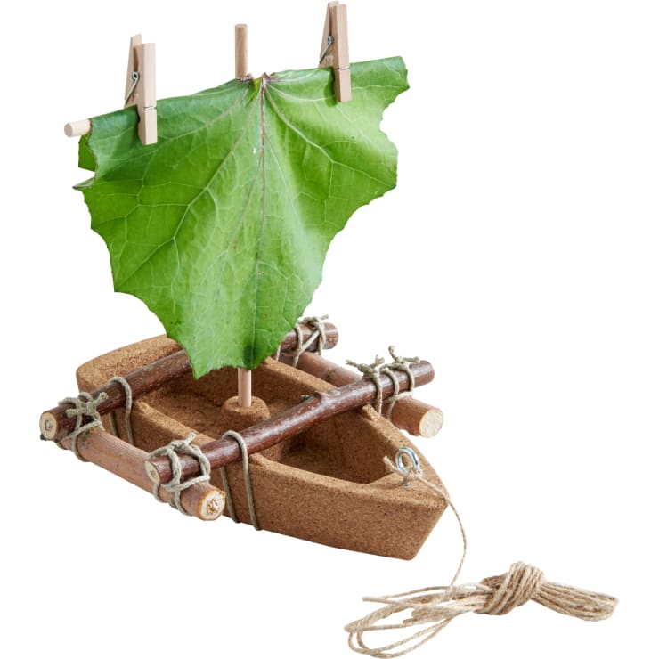 Ein kleines handgefertigtes Boot aus Kork, Zweigen und Schnur, mit einem großen grünen Blatt als Segel, das mit zwei Wäscheklammern befestigt ist, und einem Stück Seil, das vorne angebunden ist.