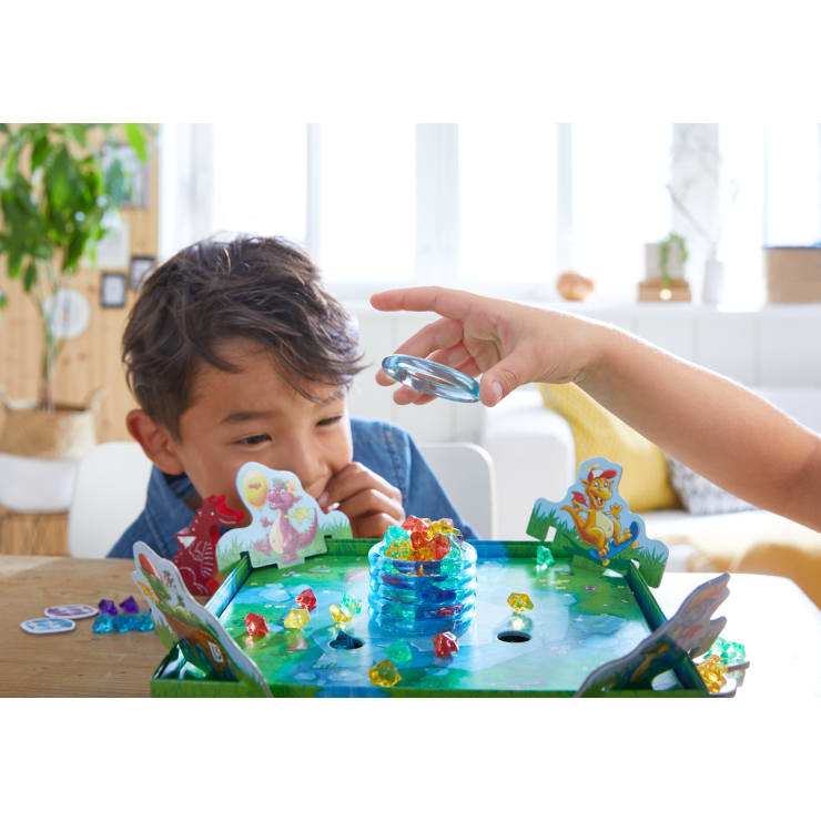 Ein kleines Kind beobachtet, wie eine andere Hand einen durchsichtigen Plastikstein auf ein buntes Brettspiel legt, das von kleinen farbigen Edelsteinen und illustrierten Tierausschnitten auf einem Tisch in einem hellen Raum umgeben ist.