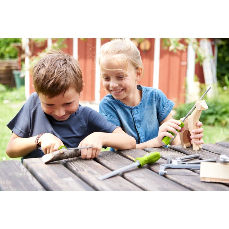 Zwei Kinder sitzen an einem Holztisch im Freien und benutzen kleine Werkzeuge, um Holzstücke zu schnitzen und zu formen. Ein Kind schnitzt konzentriert einen Zweig, während das andere lächelnd eine halbfertige Holzfigur hält.