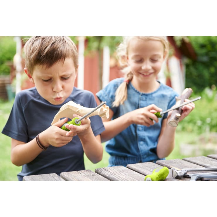 Zwei Kinder sitzen an einem Holztisch im Freien und schnitzen mit Werkzeugen an Holzstücken. Der Junge ist auf seine Arbeit konzentriert, während das Mädchen lächelt und auf ihr Projekt hinunterblickt. Die Werkzeuge liegen auf dem Tisch.