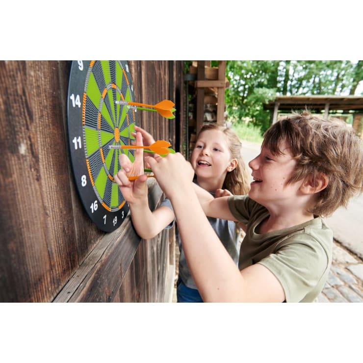 Zwei Kinder stehen neben einer Holzwand und lächeln, während sie Dartpfeile von einer Dartscheibe entfernen. Die Dartscheibe ist an der Wand befestigt und mehrere orangefarbene Dartpfeile stecken in ihr. Die Szene spielt im Freien bei Tageslicht.