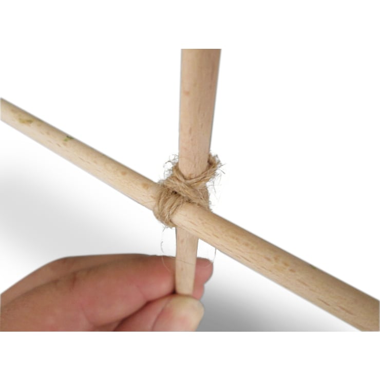 Nahaufnahme einer Hand, die zwei Holzstöcke hält, die im rechten Winkel mit Schnur zusammengebunden sind, vor einem schlichten weißen Hintergrund.