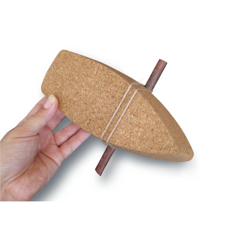 Eine Hand hält einen Korkblock in Form eines Bootes mit einem Holzstab in der Mitte, um den ein Stück Schnur gewickelt ist, vor einem weißen Hintergrund.
