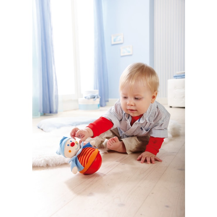 Ein Kleinkind mit hellem Haar sitzt auf einem Holzboden in einem hellen Raum und streckt die Hand aus, um ein clownförmiges Spielzeug zu berühren, das aufrecht auf einem weichen weißen Teppich sitzt.