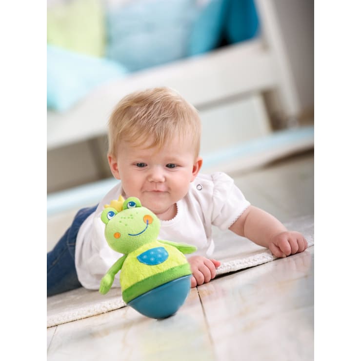 Ein Baby mit blondem Haar liegt auf einem weißen Teppich und streckt seine Hand nach einem grünen Plüschtier mit blauem Sockel in Form eines freundlichen Tieres aus. Der Hintergrund ist sanft verschwommen und suggeriert eine gemütliche Innenumgebung.