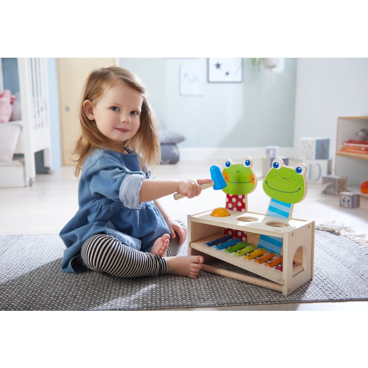 Ein kleines Kind sitzt auf einem Teppich in einem hellen Zimmer und spielt mit einem Holzspielzeug mit bunten Fröschen und einem eingebauten Xylophon, das es mit einem blauen Schlegel anschlägt.