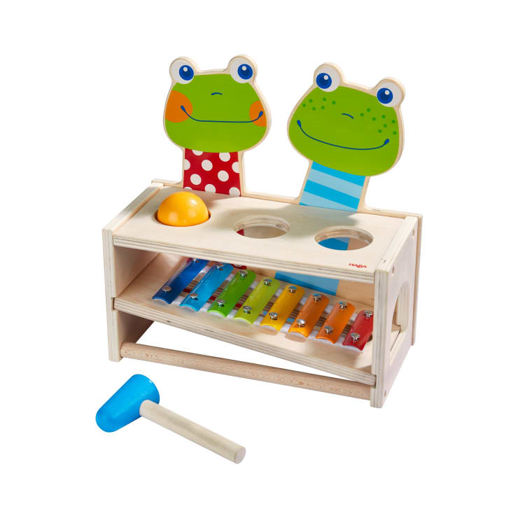 Ein Holzspielzeug mit zwei Froschgesichtern, einem gelben Ball, bunten Xylophonstäben und einem blauen Schlägel. Oben befinden sich zwei Löcher für den Ball, und das Xylophon ist in den unteren Teil der Struktur integriert.