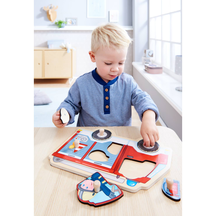 Ein kleines Kind mit blondem Haar sitzt an einem Tisch und spielt mit einem Holzpuzzle, das ein rotes Feuerwehrauto zeigt. Das Kind setzt ein Teil in das Puzzle ein, während es ein anderes Teil in der anderen Hand hält.