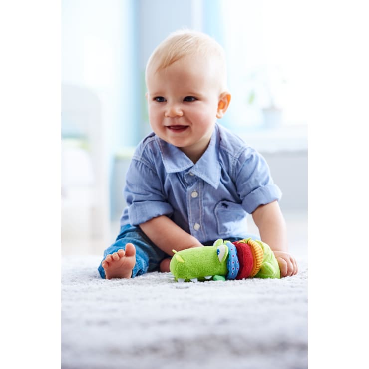 Ein Baby mit blondem Haar, blauem Hemd und Jeans sitzt auf einem hellen Teppich im Haus, lächelt und greift nach einer bunten Stoffraupe.