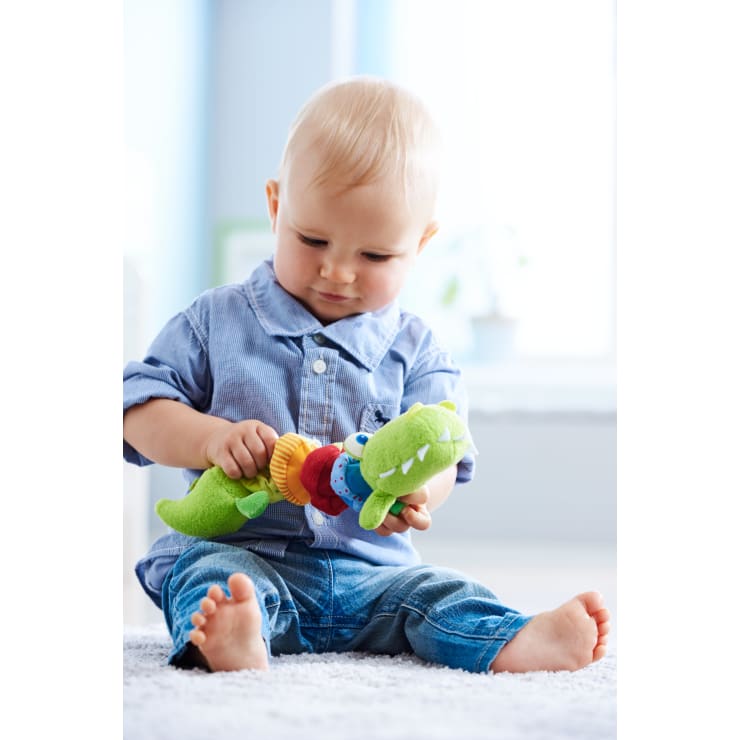 Ein Baby sitzt auf einem weißen Teppich, trägt ein blaues Hemd und Jeans, schaut nach unten und spielt mit einem grünen Plüschkrokodil. Das Zimmer ist hell erleuchtet, die Wände im Hintergrund sind hellblau.