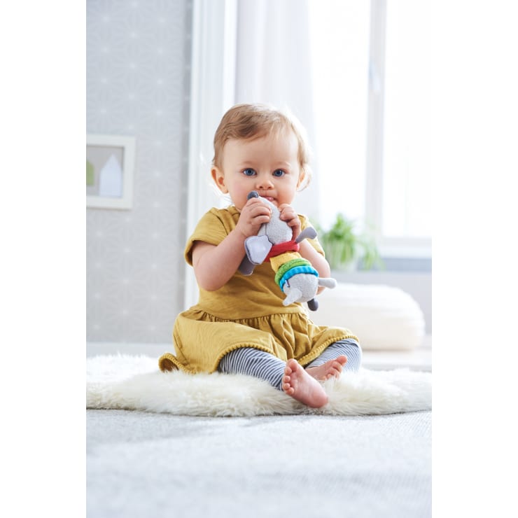 Ein Baby in einem gelben Kleid und gestreiften Leggings sitzt auf einem flauschigen Teppich, hält ein weiches, buntes Spielzeug in der Hand und kaut darauf herum. Das Zimmer ist hell mit einem weichen Hintergrund und einem Fenster, das natürliches Licht hereinlässt.