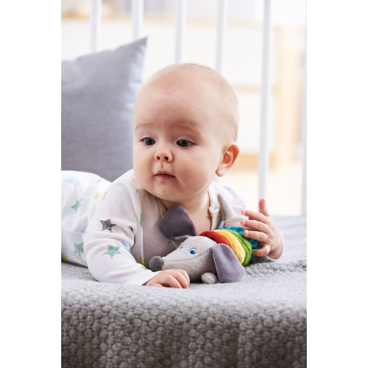 Ein Baby liegt auf einer grauen Decke, trägt einen weißen Strampler mit Sternenmuster und hält ein Plüschtier in Form eines Elefanten mit einem regenbogenfarbenen Hemd. Im Hintergrund sind ein weißes Kinderbett und ein Kissen zu sehen.