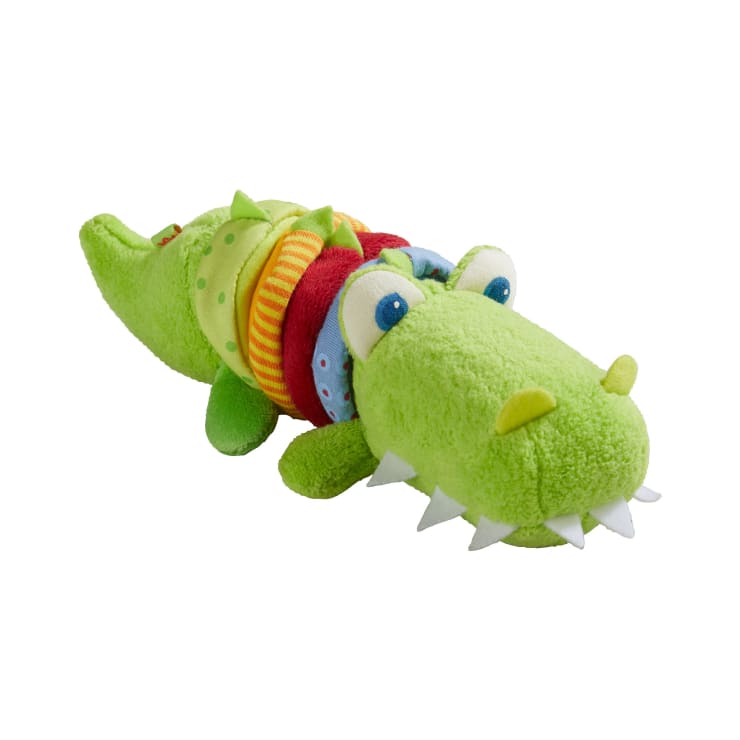 Ein grüner Plüsch-Alligator mit großen Cartoon-Augen, weißen Filzzähnen und bunten Stoffringen um den Körper auf weißem Hintergrund.
