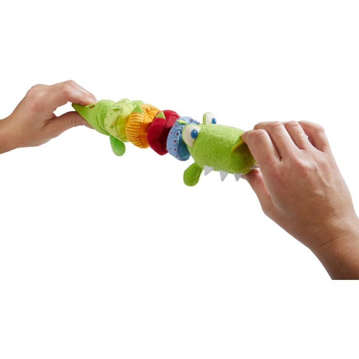 Zwei Hände halten einen weichen, grünen Plüschalligator mit bunten, abnehmbaren Ringen am Körper. Das Spielzeug hat Kulleraugen, weiße Filzzähne und verschiedene strukturierte Stoffringe. Der Hintergrund ist weiß.