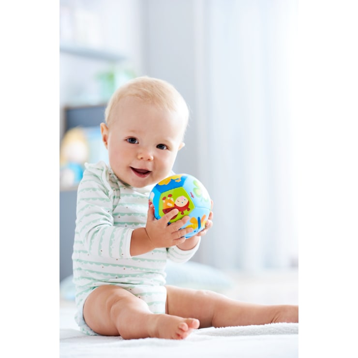 Ein Baby mit blondem Haar sitzt im Haus auf dem Boden, trägt einen hellen Strampler mit grünen Streifen und Punkten und hält einen bunten Spielzeugball mit Tiermotiven.