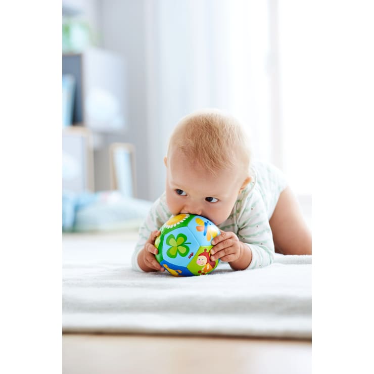 Ein Baby in einem hellen Strampler liegt auf einem weichen, weißen Teppich und hält einen bunten Spielzeugball mit Tier- und Kleemotiven in der Hand und beißt hinein. Der Hintergrund ist sanft verschwommen und zeigt ein helles, gemütliches Zimmer.