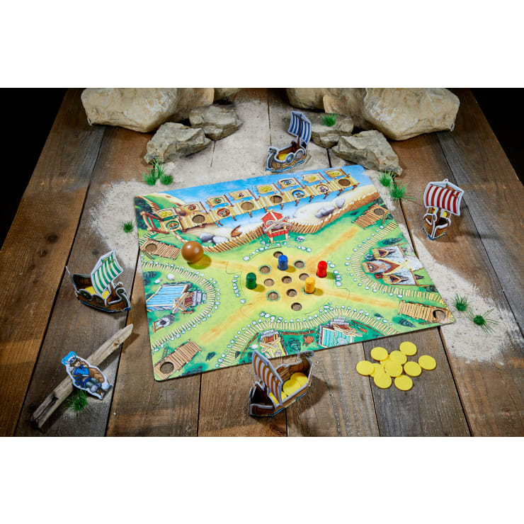 Ein farbenfrohes Brettspiel mit Wikingerfiguren, vier kleinen Modellschiffen, gelben Spielsteinen und vier farbigen Spielfiguren auf einem Brett, das auf einem Holztisch mit Steinen und Grashalmen als Dekoration angeordnet ist.