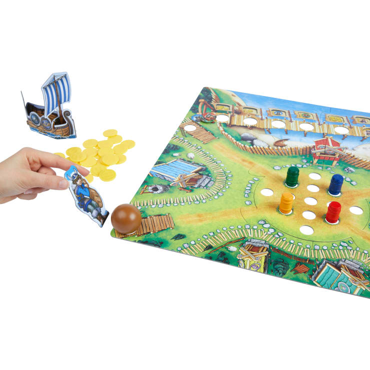 Eine Hand legt ein Wikinger-Figurenplättchen an den Rand eines Brettspiels mit einem grünen Pfad, farbigen Spielsteinen in Löchern, einem braunen Ball, gelben Münzplättchen und einem Wikingerschiff-Plättchen.