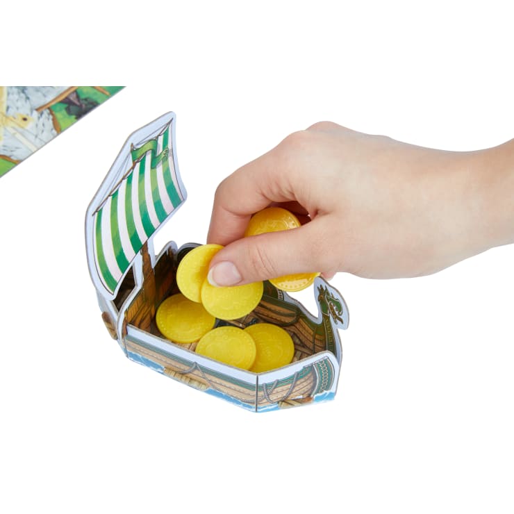 Eine Hand greift in ein kleines Pappboot, das mit gelben Plastikmünzen gefüllt ist. Das Boot hat ein grün-weiß gestreiftes Segel und ist Teil eines Brettspiels.