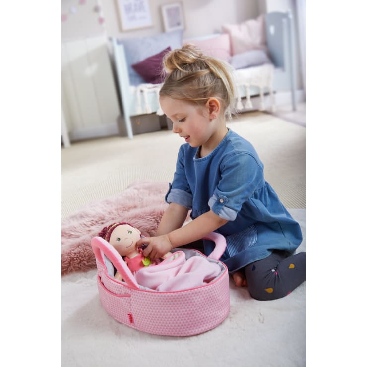 Ein junges Mädchen in einem blauen Kleid sitzt auf einem rosa Teppich und legt eine Puppe in einen rosa Tragekorb. Die Szene spielt in einem gemütlichen, sanft dekorierten Zimmer mit einem Kinderbett und einem Bett im Hintergrund.