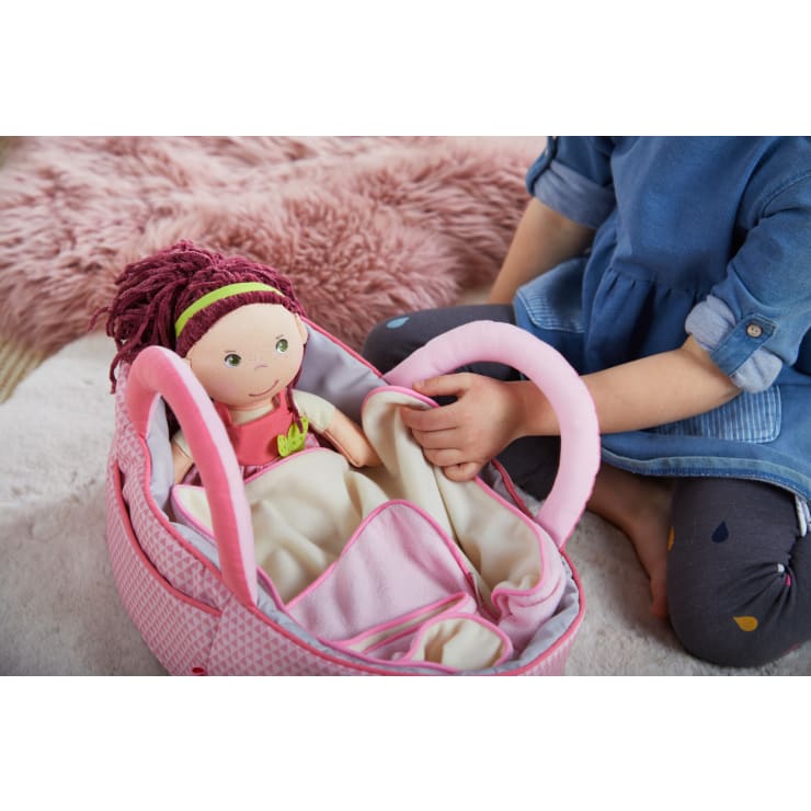 Ein Kind sitzt auf dem Boden neben einem weichen rosa Teppich und hält eine rosa Spielzeugtrage mit einer Stoffpuppe darin. Die Puppe hat Garnhaare und ist in eine Decke eingewickelt. Nur die Hände und der Unterkörper des Kindes sind sichtbar.