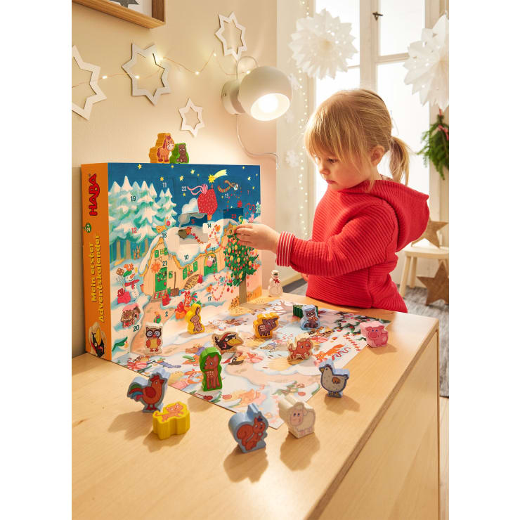 Ein kleines Kind in einem roten Pullover spielt mit einem bunten Adventskalender auf einem Holztisch, auf dem verschiedene kleine Tierfiguren verstreut sind. Der Raum ist mit Papiersternen geschmückt und wird von natürlichem Licht beleuchtet.