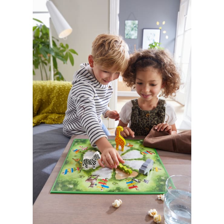 Zwei kleine Kinder spielen ein Brettspiel mit Tierfiguren, darunter ein Zebra, eine Giraffe und ein Krokodil, auf einem grünen Brett. Ein Glas Wasser und verschüttetes Popcorn stehen auf dem Tisch. Die Szene spielt sich in einem hellen Wohnzimmer ab.