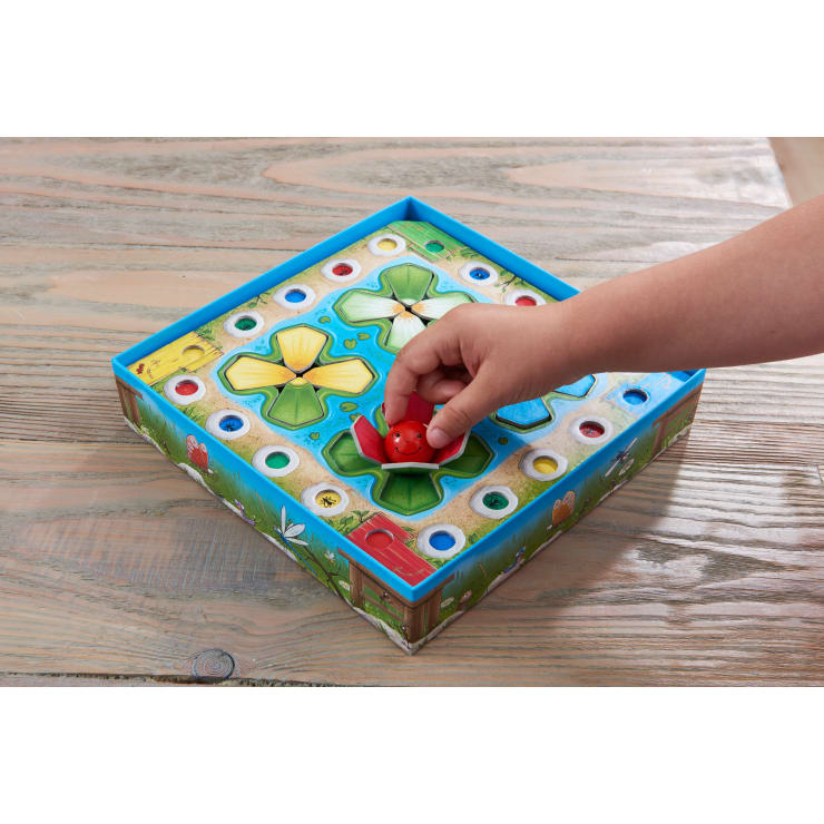 Eine Kinderhand setzt eine rote Spielfigur auf ein buntes Spielbrett mit vier Feldern und kreisförmigen Löchern, das auf einem Holztisch liegt.