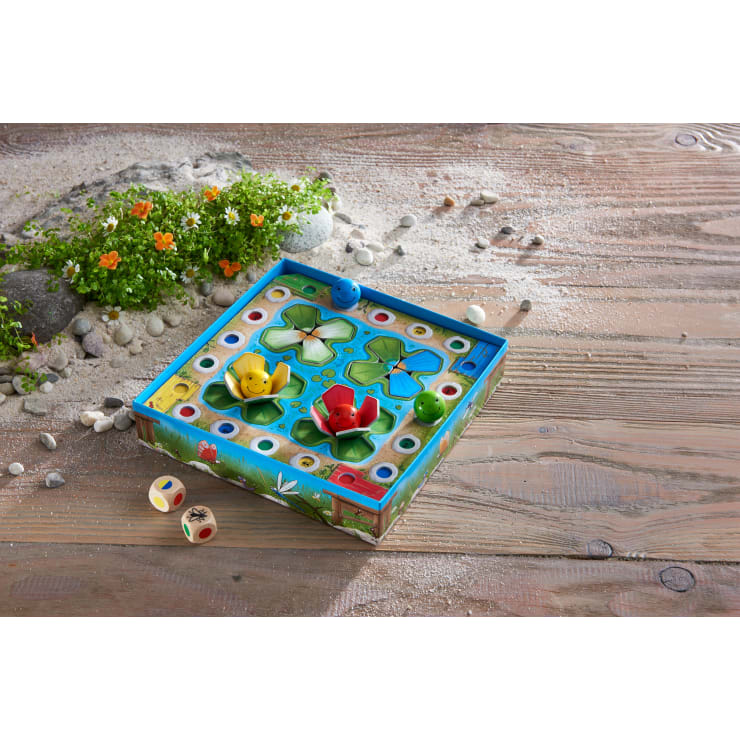 Ein farbenfrohes Brettspiel mit zwei Froschfiguren, einer gelben und einer roten, liegt auf einer hölzernen Oberfläche mit verstreuten Kieselsteinen, drei Würfeln und einem Beet mit kleinen orangefarbenen und weißen Blumen an der Seite.