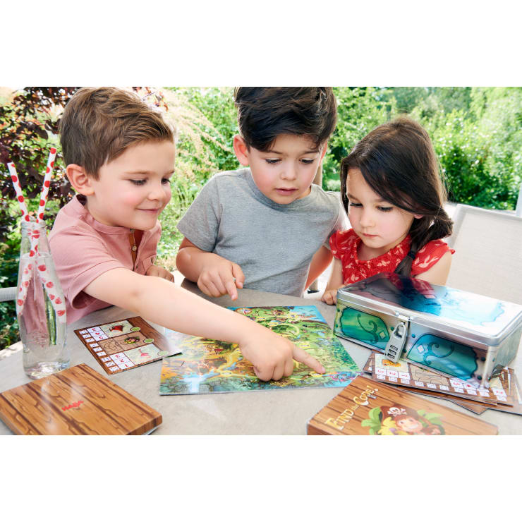 Drei Kinder sitzen an einem Tisch im Freien und sehen sich ein bunt illustriertes Brettspiel an. Zwei Jungen und ein Mädchen beschäftigen sich mit dem Spiel, während eine Lunchbox aus Metall und gemusterte Getränke auf dem Tisch stehen. Es ist Tageszeit.