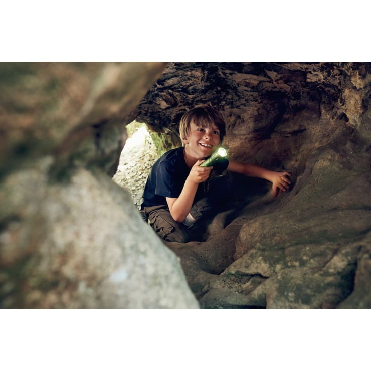 Ein Junge mit einer Taschenlampe in der Hand hockt in einer Felsenhöhle, schaut nach vorne und lächelt. Das Sonnenlicht fällt durch den Eingang und beleuchtet einen Teil der Höhlenwände und das Gesicht des Jungen.