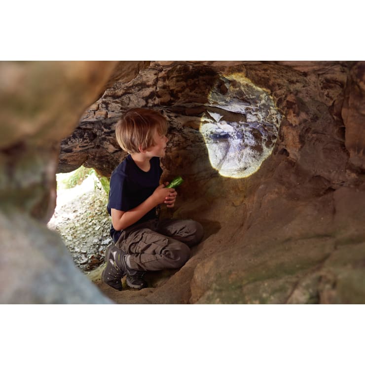 Ein kleiner Junge kniet in einer kleinen Höhle oder Felsmulde, hält eine Taschenlampe in der Hand und leuchtet damit auf die Höhlenwand. Er trägt ein marineblaues T-Shirt und eine Cargohose und schaut auf die beleuchtete Stelle auf den Felsen.