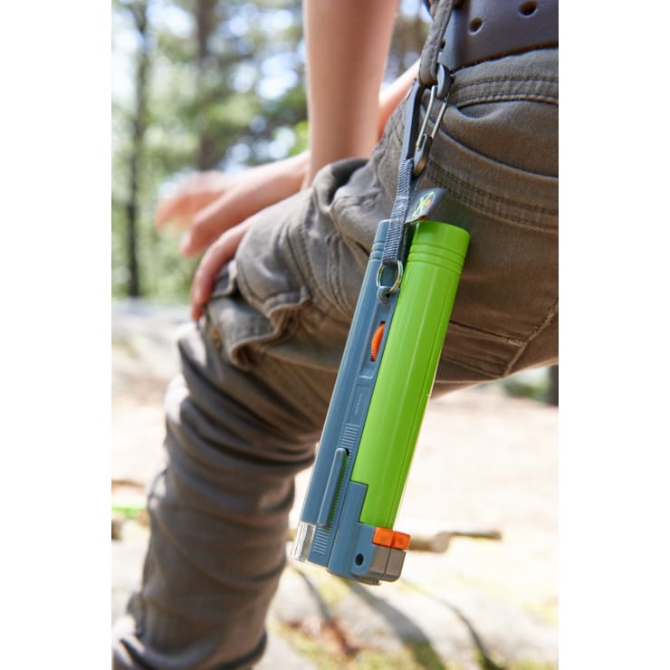 Eine Person in einer grauen Hose sitzt im Freien und hat eine grün-blaue Taschenlampe an der Gürtelschlaufe befestigt. Im unscharfen Hintergrund sind Bäume und Felsen zu sehen.