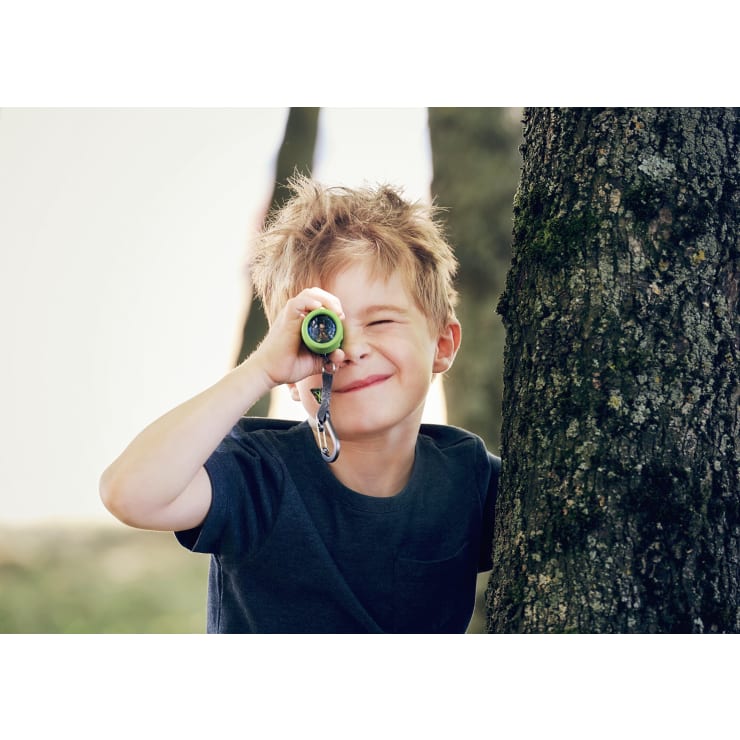 Ein kleiner Junge mit unordentlichem blondem Haar steht an einem Baum, hält ein kleines grünes Spielzeugteleskop an sein Auge und lächelt. Er trägt ein dunkelblaues T-Shirt und befindet sich im Freien, mit verschwommenen Bäumen im Hintergrund.