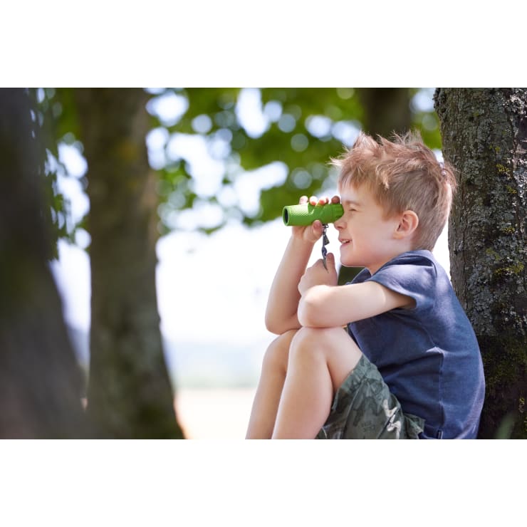 Ein kleiner Junge sitzt im Freien an einem Baum, hält ein grünes Spielzeugteleskop an sein Auge und schaut in die Ferne. Er trägt ein blaues Hemd und Shorts in Tarnfarben. Der Hintergrund zeigt verschwommene Bäume und Sonnenlicht.