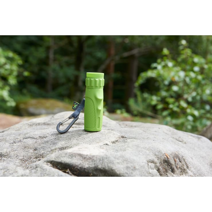 Eine kleine grüne Taschenlampe mit einem grauen Karabinerhaken sitzt aufrecht auf einem großen Felsen im Freien, mit grünem Laub und Bäumen im verschwommenen Hintergrund.