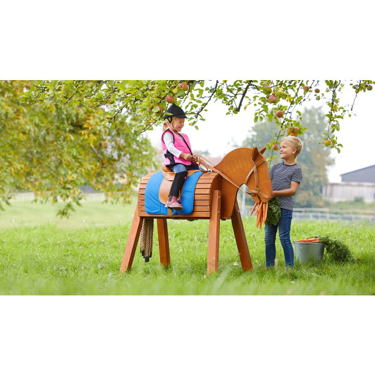 Zwei Kinder sitzen draußen unter einem Baum; eines sitzt auf einem hölzernen Spielzeugpferd und hält die Zügel in der Hand, während das andere in der Nähe steht und ein Bündel Möhren neben einem mit Gras und Möhren gefüllten Metalleimer hält.