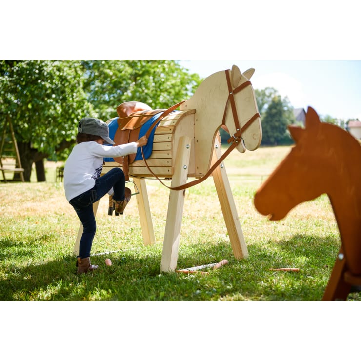 Ein Kind mit Hut und Stiefeln klettert auf ein hölzernes Spielzeugpferd im Freien auf einer Wiese, mit einem anderen Holzpferd im Vordergrund und Bäumen im Hintergrund.