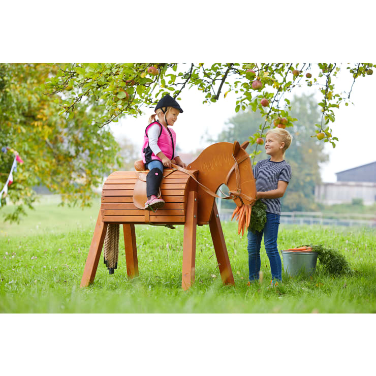 Ein Mädchen in Reitkleidung sitzt im Freien auf einem hölzernen Spielzeugpferd, während ein Junge neben ihr steht und ein Bündel Karotten in der Hand hält. Ein Eimer mit weiteren Karotten steht in der Nähe unter einem Apfelbaum.